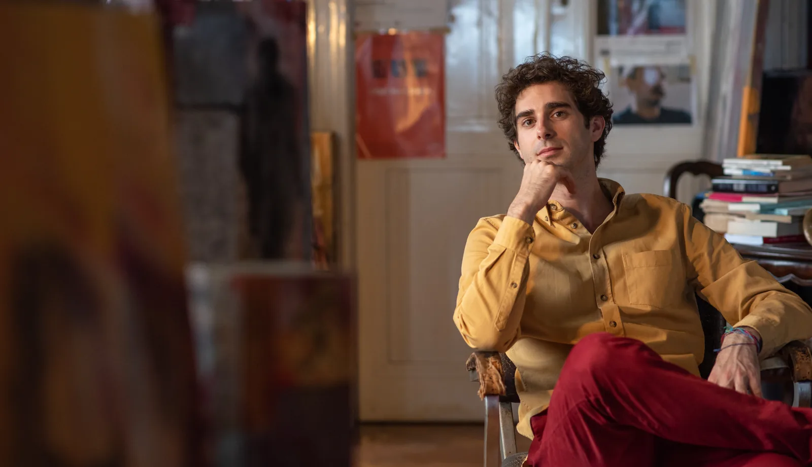 Luca Sacher seduto in poltrona nello studio, camicia gialla e pantaloni rossi, fra libri e quadri