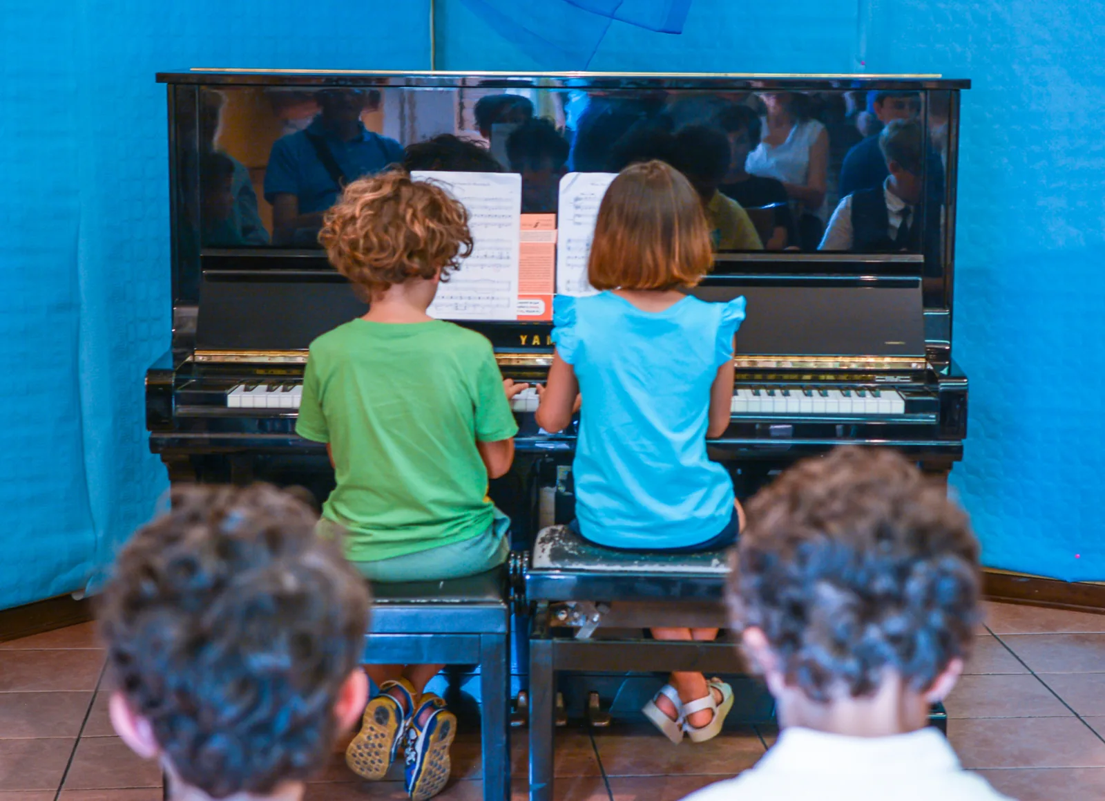 Due bambini suonano a quattro mani sul pianoforte verticale, davanti al pubblico del saggio